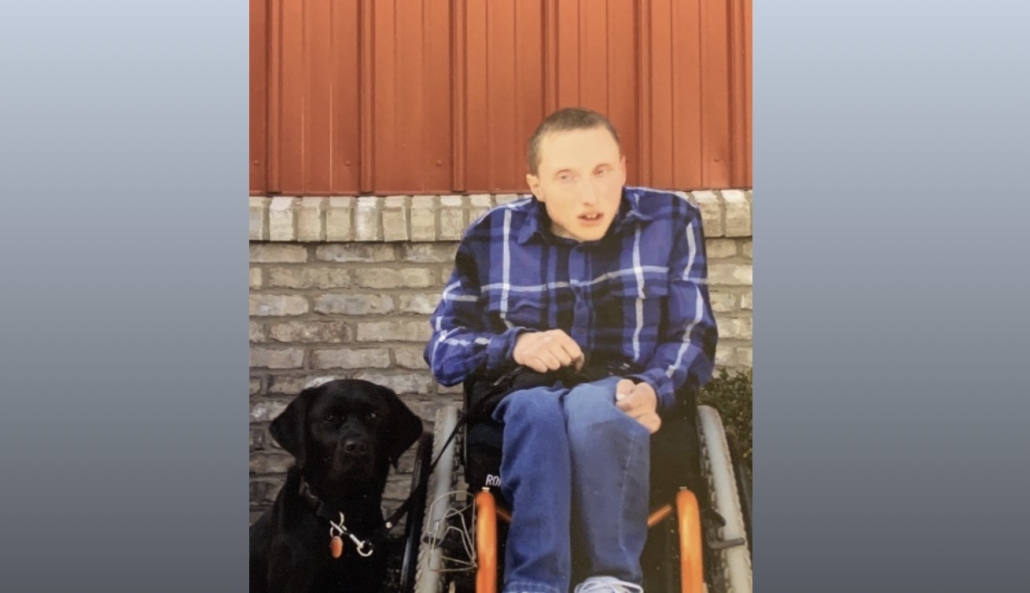 Fostoria News Young man in wheelchair with black dog outside on brick wall background.