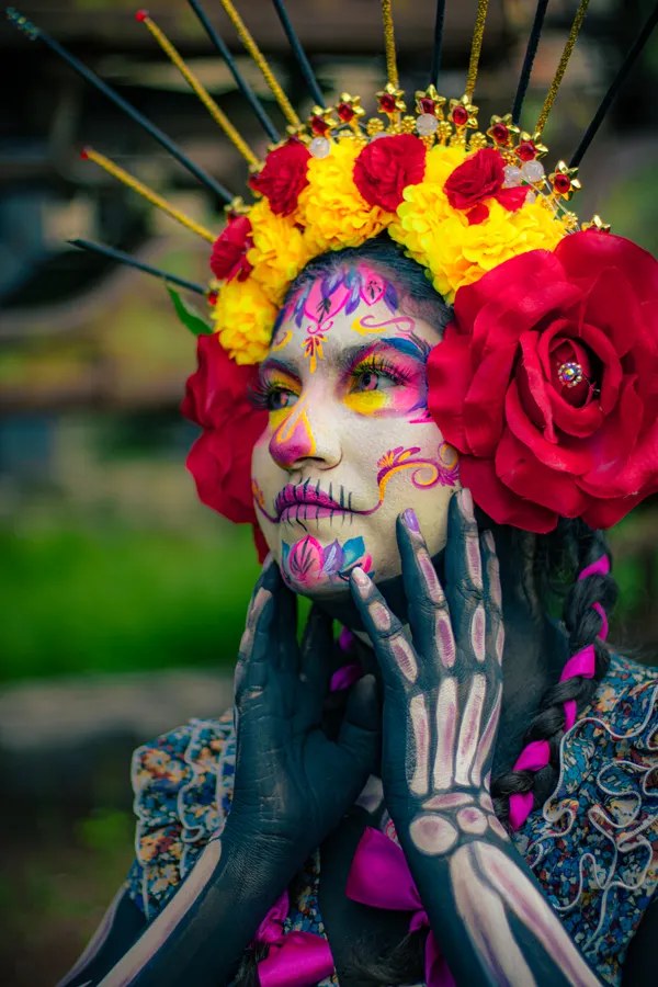 Fostoria News: Woman in colorful Catrina face paint wearing a crown of marigolds and red roses, with skeleton designs painted on her hands