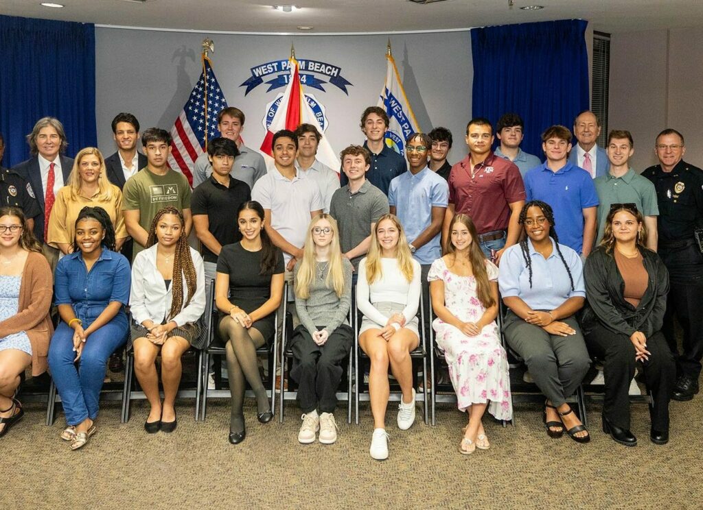 A group of people posing together indoors. The group consists of young adults seated in the front, with adults standing behind them, including several individuals in police uniforms. Flags and a banner are displayed in the background.