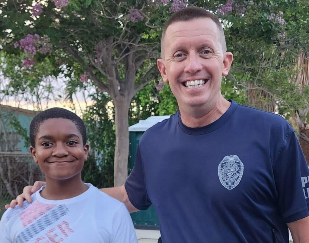 A man in a navy blue police uniform stands smiling with his arm around a boy wearing a white T-shirt with red text. They are outdoors in front of a tree with purple flowers and a fence in the background.