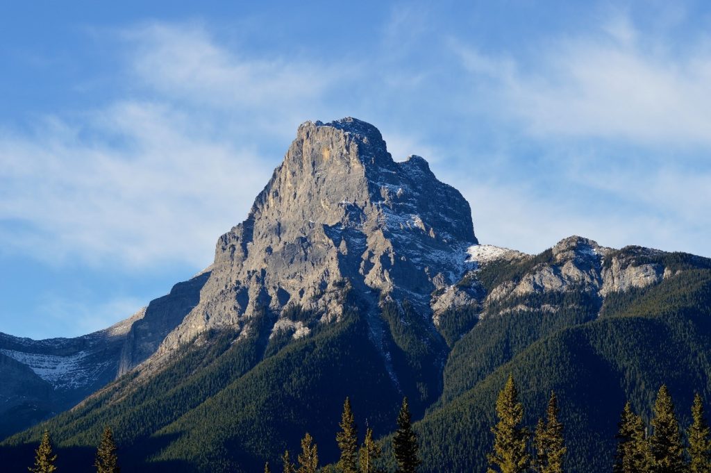 A rugged mountain peak rises above a forested hillside under a blue sky with wispy clouds.