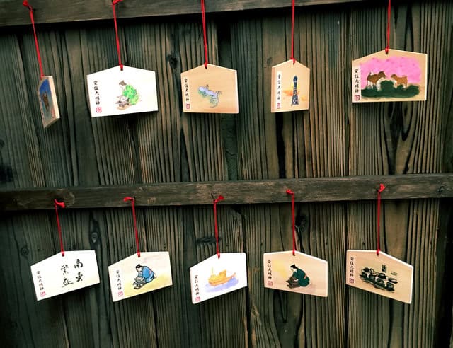 Ten small, rectangular wooden prayer plaques, or ema, featuring various hand-painted illustrations, hanging from red strings on a wooden wall.