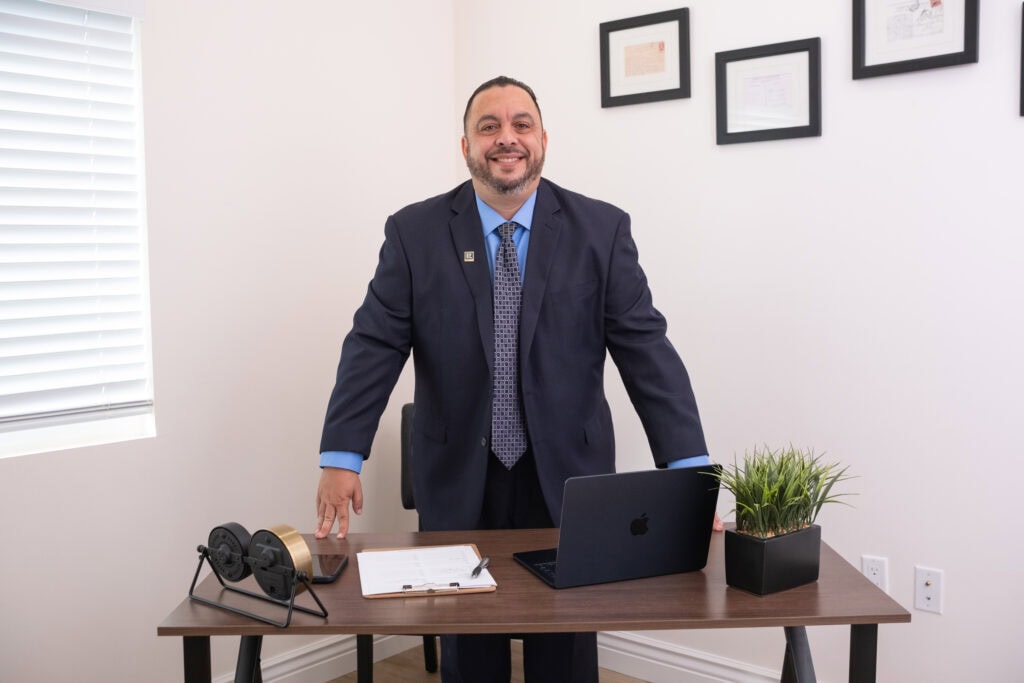 Professional businessman Tony Tadros in office, smiling, standing at desk with laptop, plant, and documents.