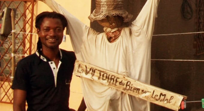 Jeune homme souriant à côté d'une statue traditionnelle togolaise.