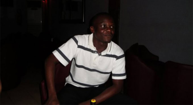 Homme togolais assis dans un intérieur sombre, portant un polo rayé blanc et noir.