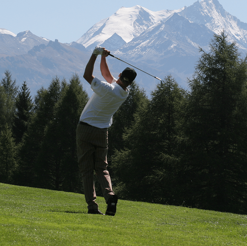 A man takes a powerful golf swing on a scenic course, silhouetted against majestic mountains and a line of pine trees.