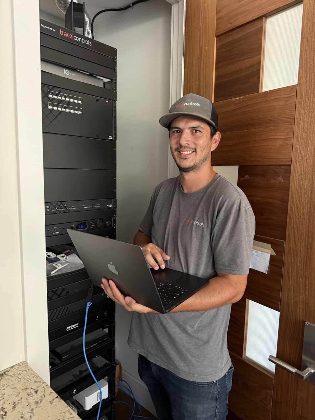 Adam, a smart home integrator, smiling with a laptop in front of an equipment rack of networking and audio video gear in a client's home