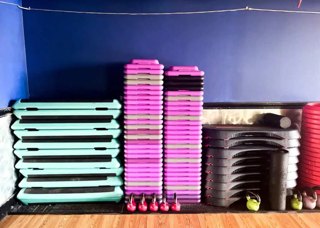Stacked step platforms and kettlebells organized against a blue wall in the Triangle Fitness group exercise studio.