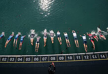 Group of athletes swimming in open water at the start of a triathlon race, with a timing mat and official overseeing the event.