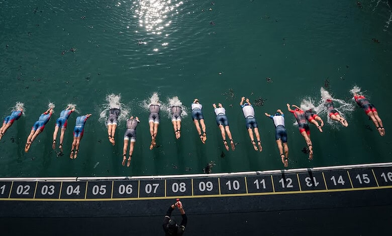 Group of athletes swimming in open water at the start of a triathlon race, with a timing mat and official overseeing the event.