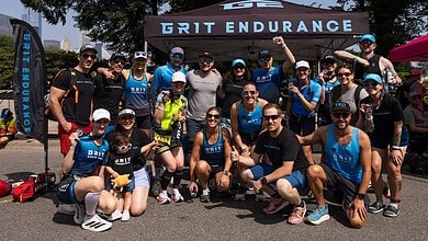 Group of athletes posing for a photo under a GT endurance tent at a race event, wearing blue and black triathlon apparel, sunglasses and caps, sunny day.