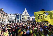 Large crowd of cyclists and spectators at the Tour de France start line in a city square, with a prominent yellow Tour de France flag and historic buildings in the background.