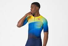 Male triathlon athlete wearing a colourful cycling suit with yellow, blue, and green shades, standing in a studio with a white background.