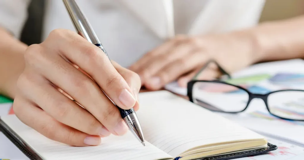A person's hand writing in a notebook with a pen and glasses | tuitioncentre sg