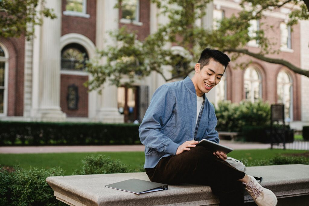 Asian male using tablet while resting on bench in University backyard | Tuition Centre SG