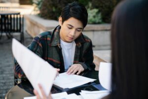 A focused young man in a plaid jacket reviews documents outdoors at a table. Another person's blurred back is in the foreground | Tuition Centre SG