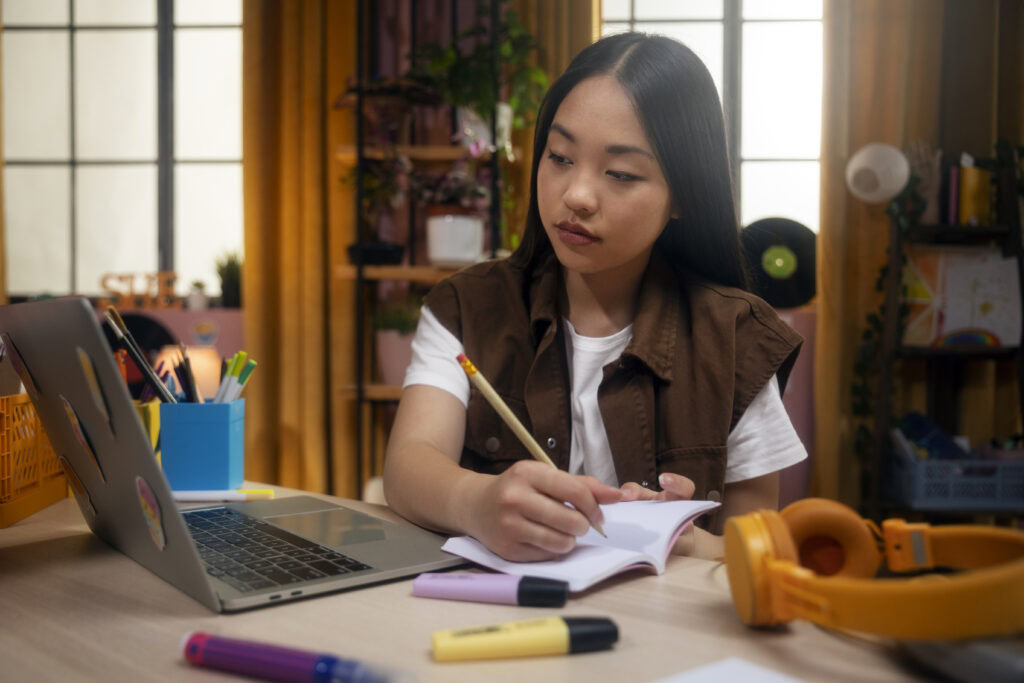 Teen girl taking notes while studying at a desk with a laptop, notebooks, and stationery in a cozy room. | TCS