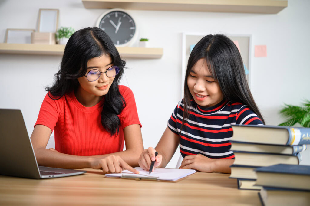 Two girls studying together at a desk with books, laptop, and notepad, smiling and writing notes. | TCS