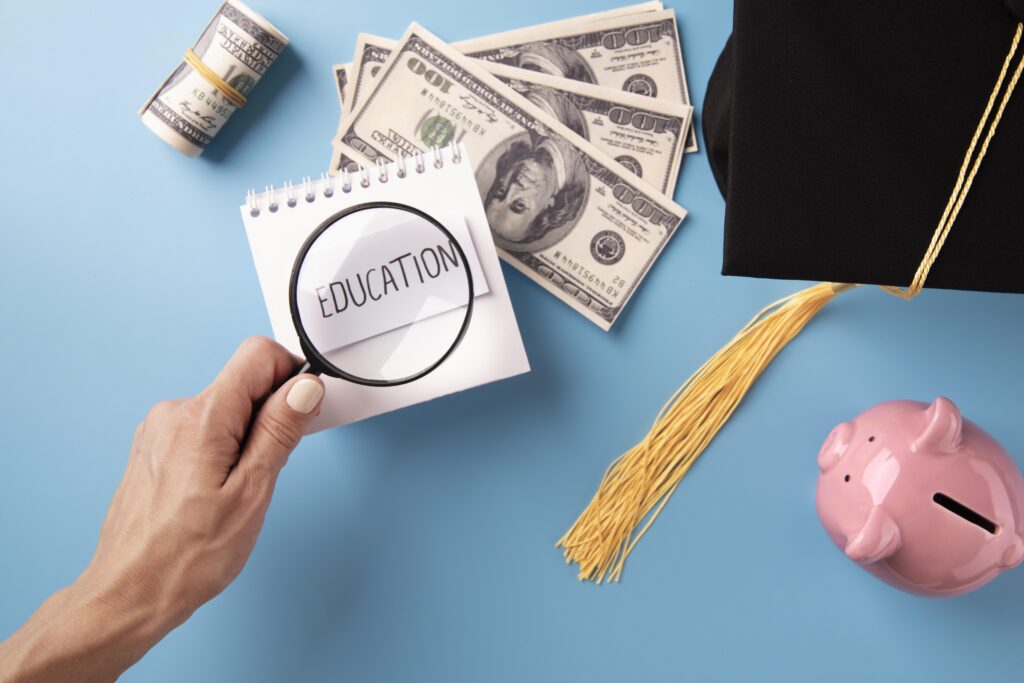 A hand holds a magnifying glass over a notepad labeled "Education" near cash, a piggy bank, and a graduation cap. | TCS