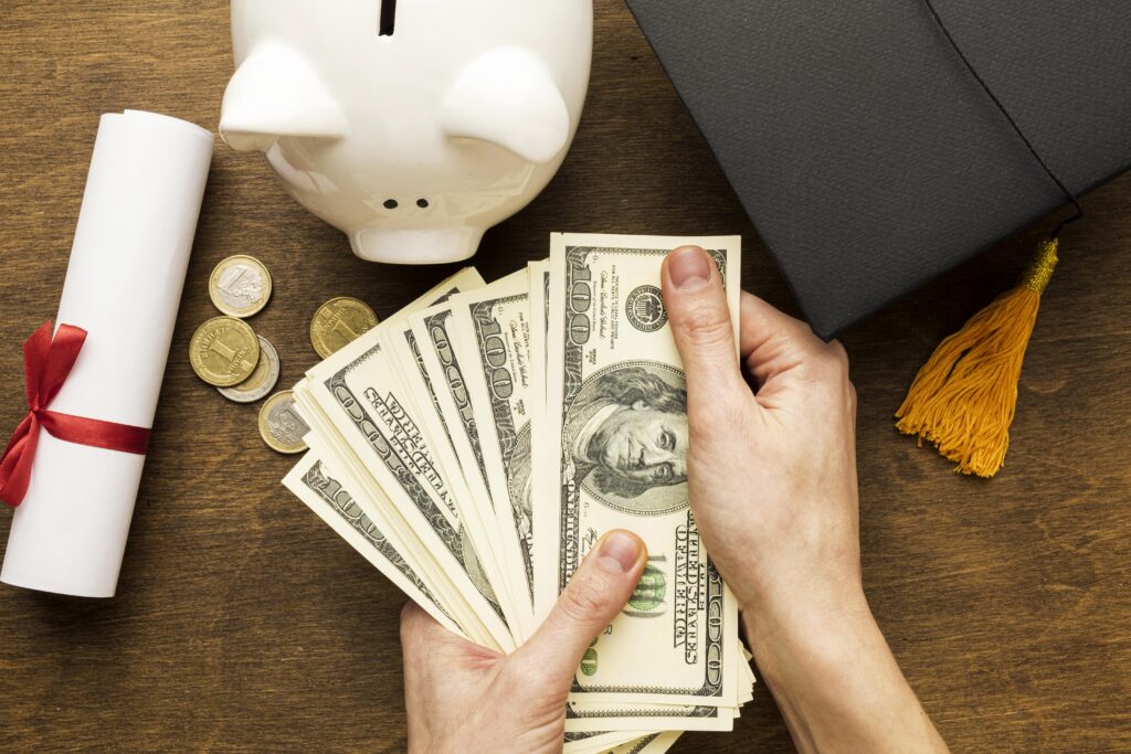 Hands holding dollar bills near a piggy bank, coins, graduation cap, and a diploma on a wooden table. | TCS