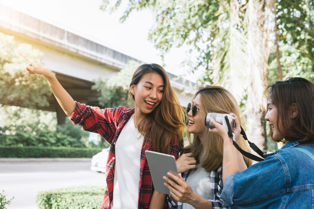Three young women outdoors, one holding a tablet, another filming with a camcorder, and one pointing ahead, all smiling. | TCS