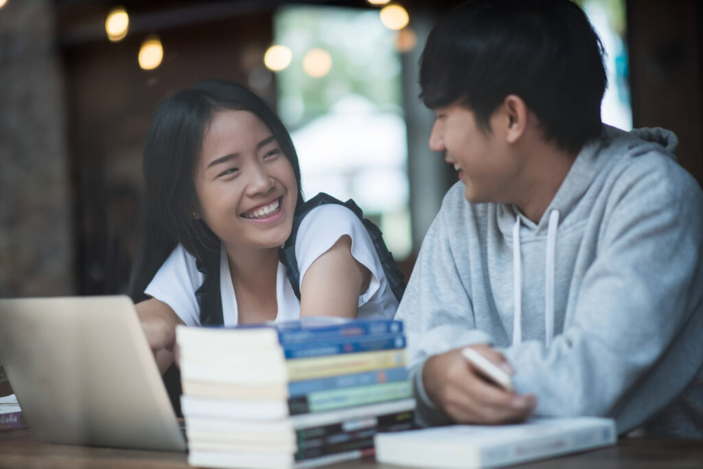 Two students sit at a table with books and a laptop, smiling and talking to each other in a cozy setting. | TCS