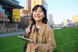 Smiling young woman with a tablet and backpack stands outside in a city park with modern buildings behind her. | TCS