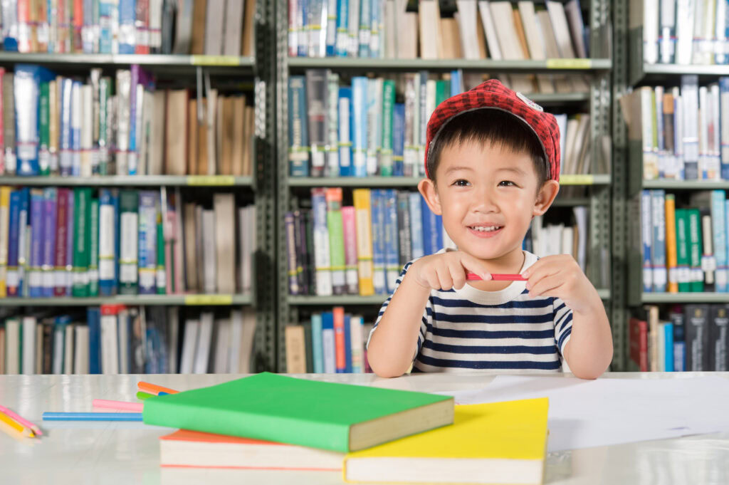 Smiling child in a striped shirt and red cap sits at a library table with books and colored pencils. | TCS