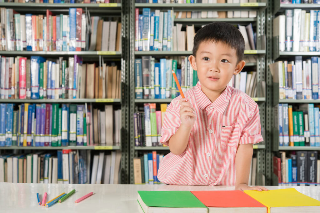 Young boy in a pink striped shirt holds a pencil, sitting at a table with colorful books in a library. | TCS