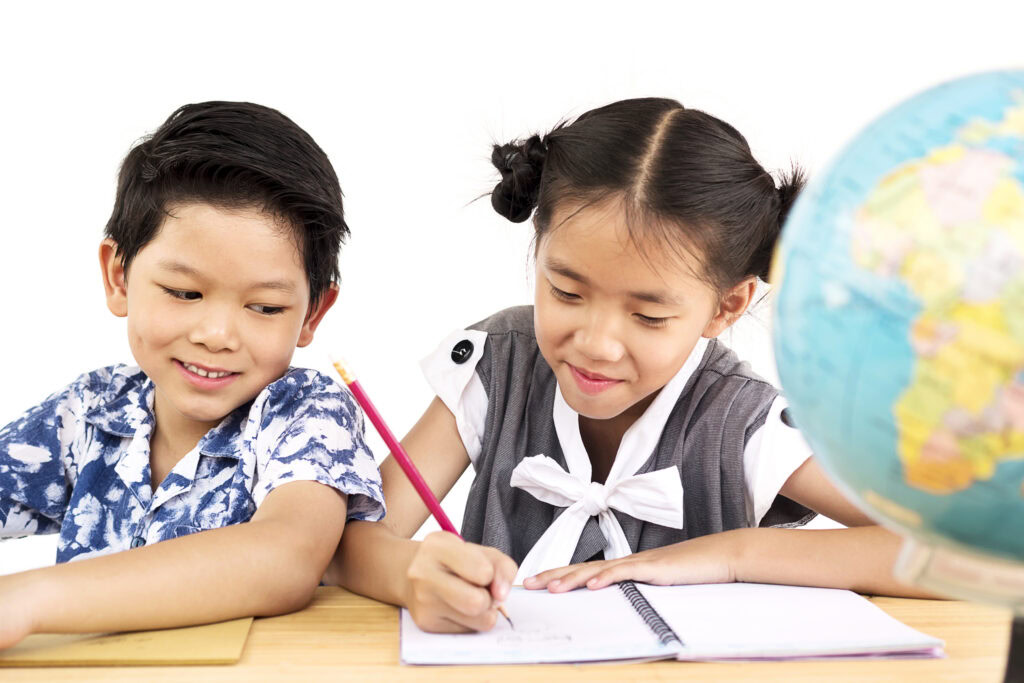 Two children sit at a desk, writing in notebooks, with a globe visible in the foreground on the right. | TCS