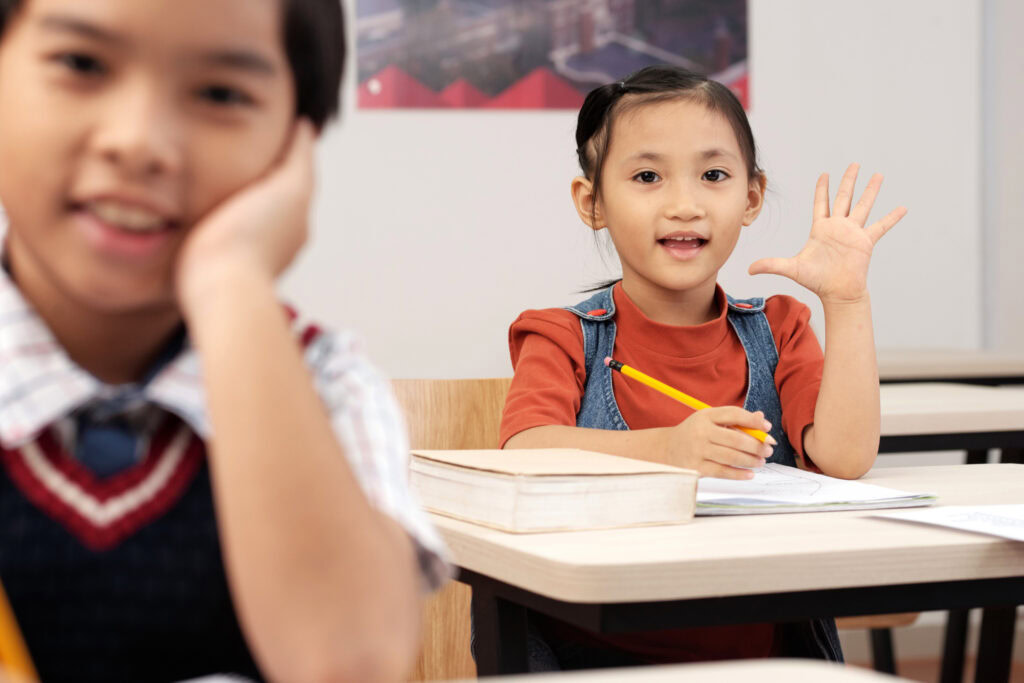 Young girl sitting at a desk in a classroom, smiling and raising her hand, while a boy sits in the foreground. | TCS