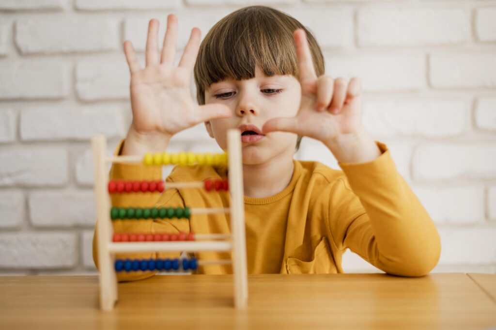 A young child in a yellow shirt counts on their fingers in front of a colorful abacus on a table. | TCS