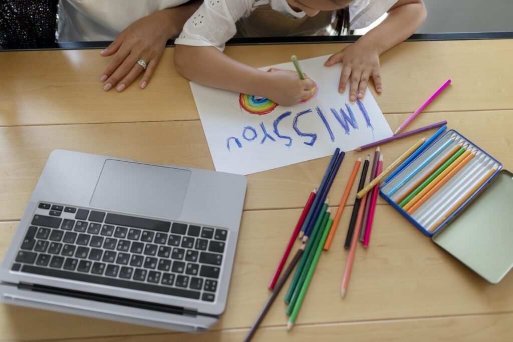 Child drawing "I MISS YOU" with colored pencils near a laptop, adult hand nearby on a wooden table. | TCS