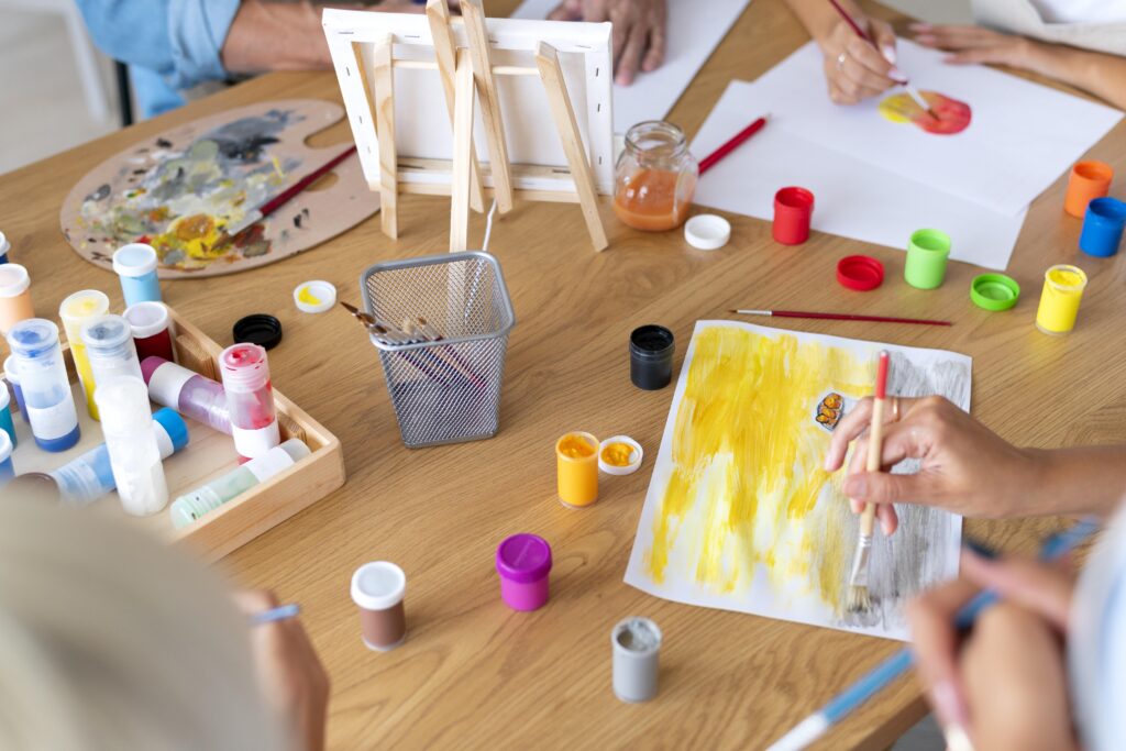 People painting with brushes and colorful paints at a table, with paper, jars of paint, and art supplies scattered around. | TCS