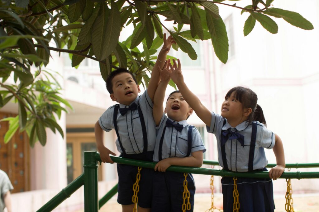 Three young children in uniforms reach up toward leaves while standing on a green playground structure. | TCS