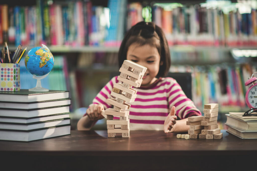 A young girl in a striped shirt plays with a tumbling wooden block tower at a library table with books and a small globe. | TCS