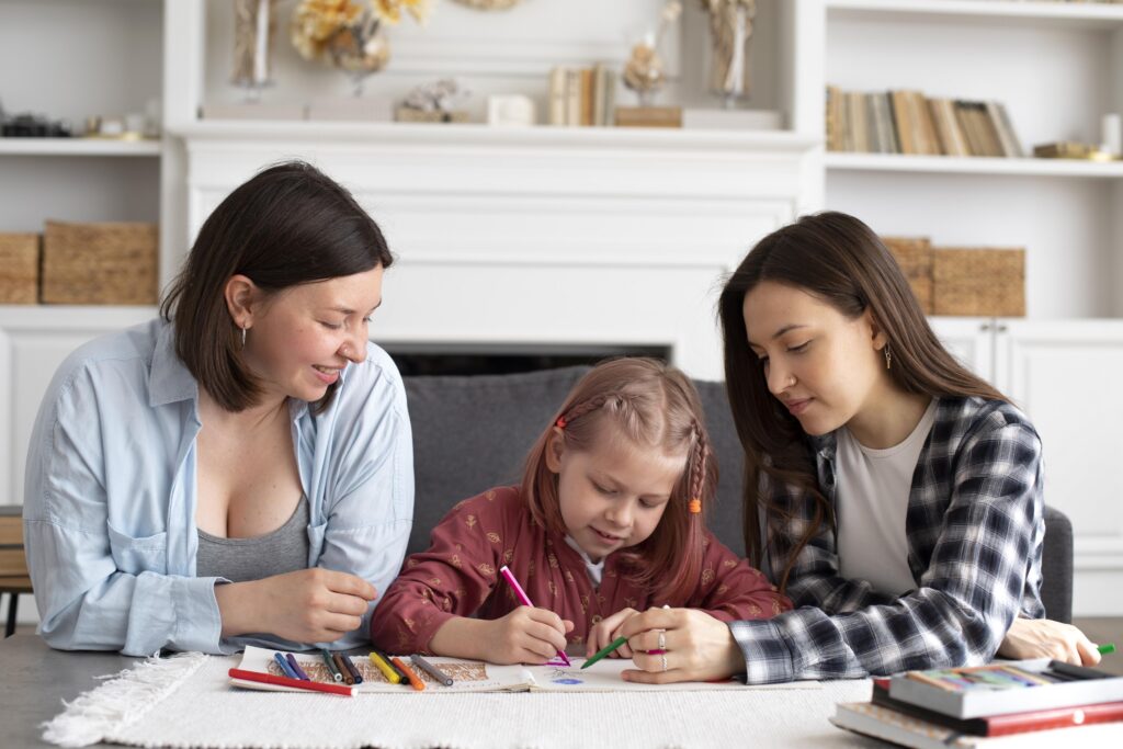Two women watch a young girl coloring with crayons at a table in a cozy living room with shelves and books in the background. | TCS
