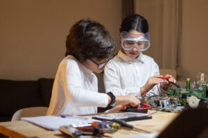 Two children work on electronics at a table, one using a tablet, the other wearing safety goggles and holding wires. | TCS
