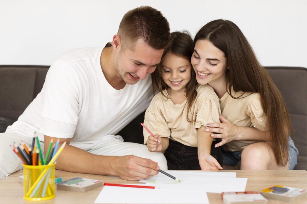 A smiling family sits together, coloring and drawing on paper at a table with colored pencils. | TCS