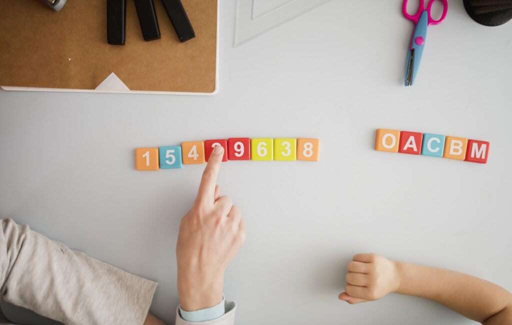 A hand points to numbered blocks arranged in sequence on a table, with lettered blocks and school supplies nearby. | TCS