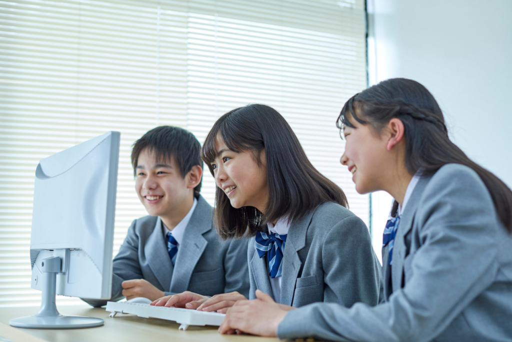 Three students in school uniforms smiling and looking at a computer monitor in a bright classroom. | TCS