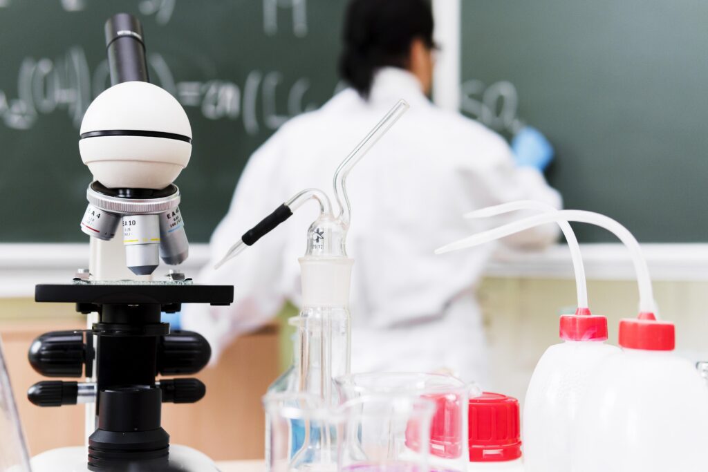 A scientist writes on a blackboard in a laboratory, with a microscope and glassware in the foreground. | TCS
