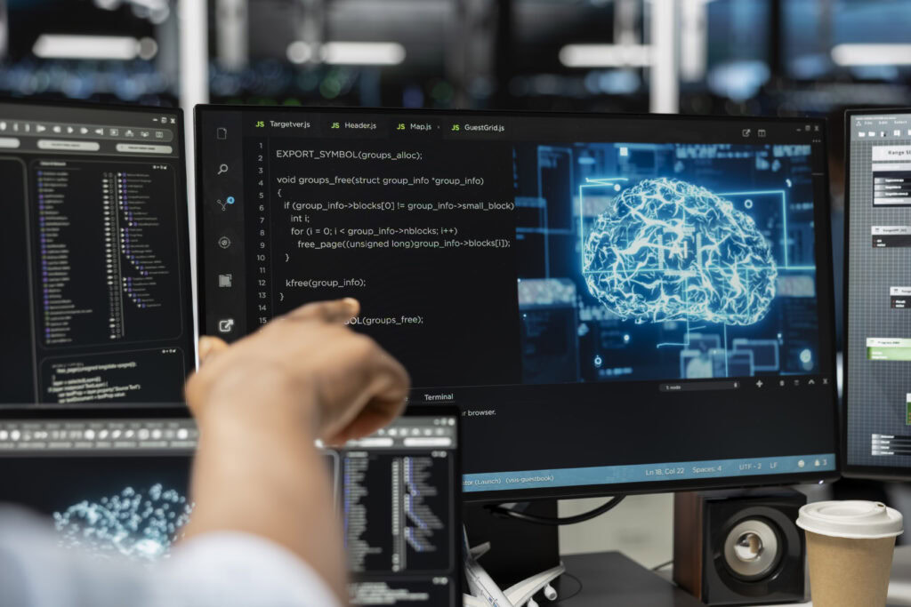 Person points at a monitor displaying AI code and a digital brain graphic in a high-tech workspace. | TCS