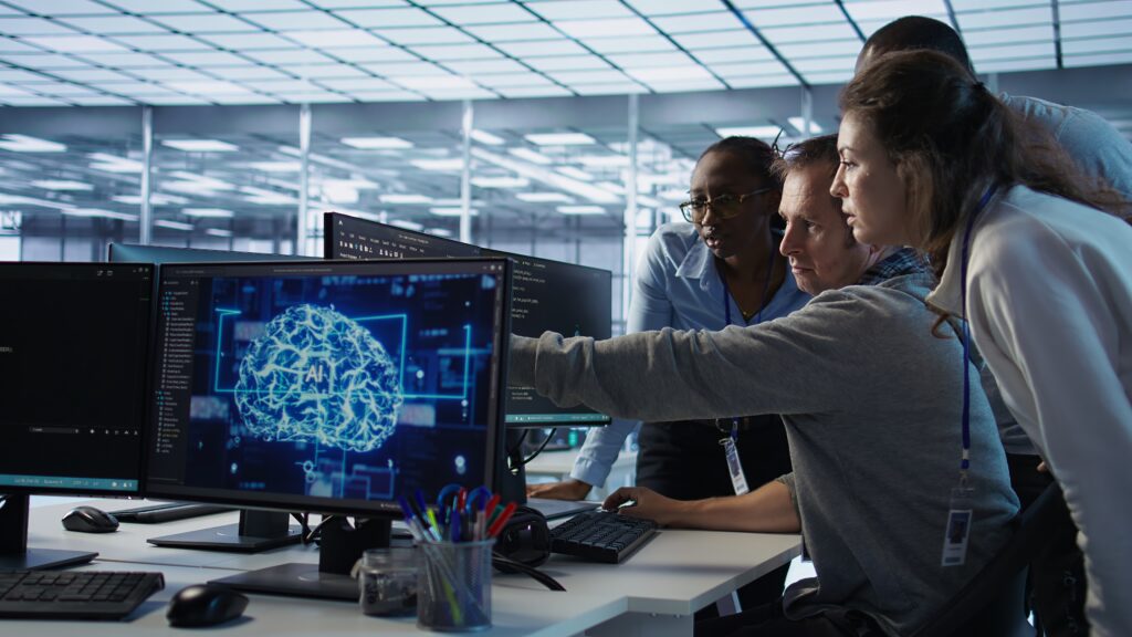 Four people collaborate at a desk, looking at a computer screen displaying a digital brain graphic in an office setting. | TCS