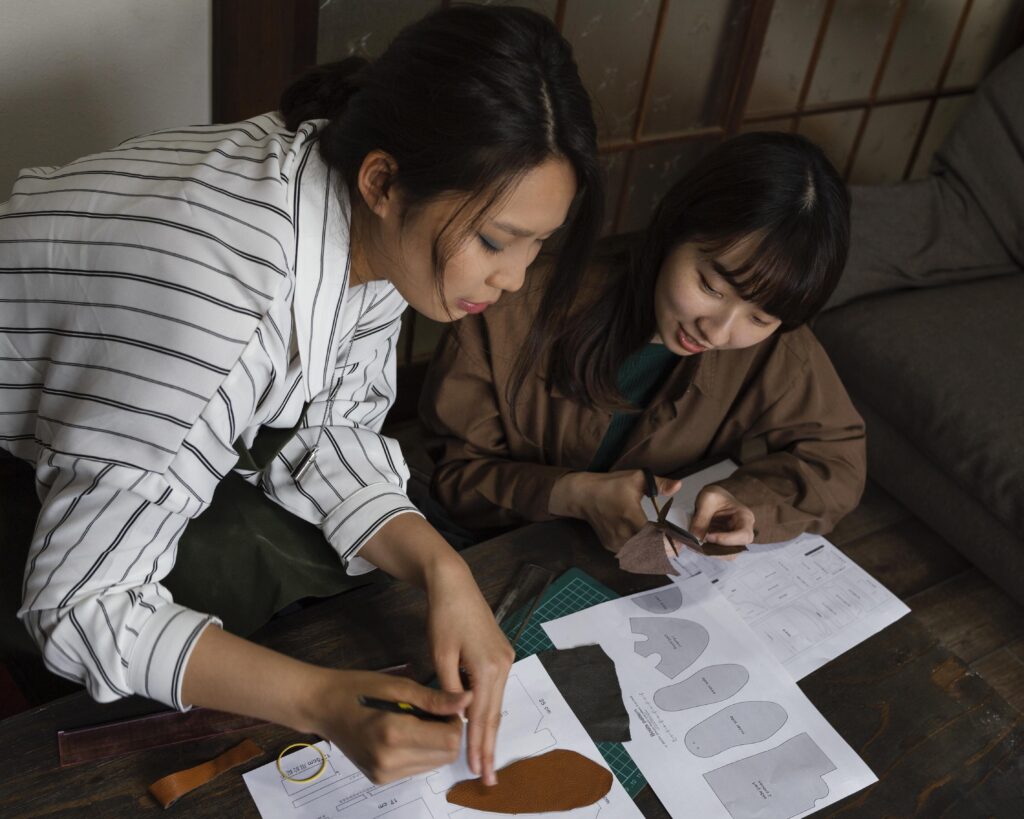 Two women working on leathercraft projects at a table, using scissors and tools with pattern papers in front of them. | TCS