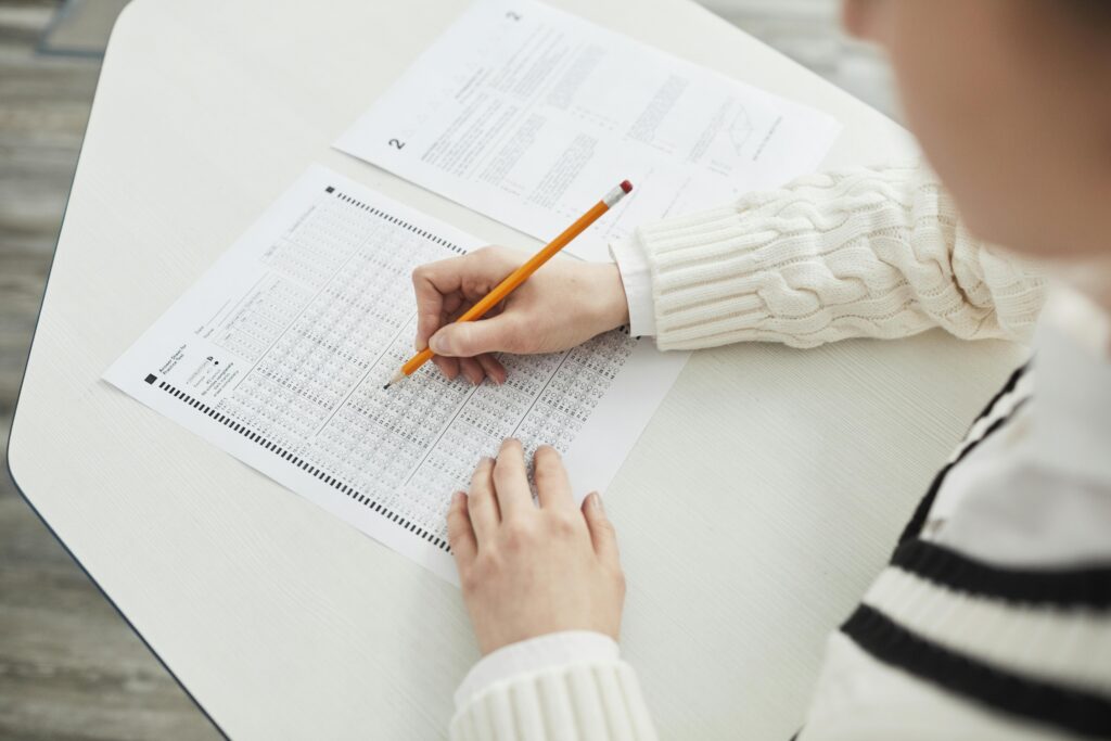 Person filling out a multiple-choice answer sheet with a pencil at a desk, wearing a white sweater. | TCS