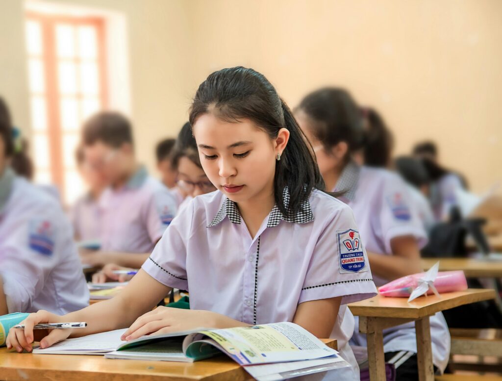 A student in uniform reads a textbook and writes at her desk in a classroom with other pupils. | TCS