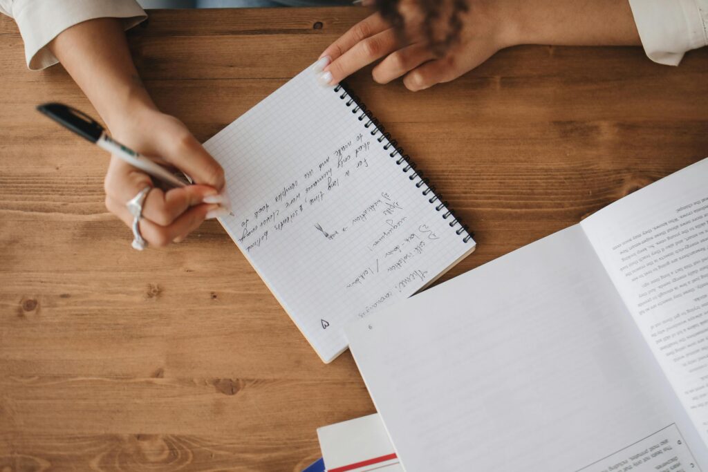 Two people at a wooden table, one writing notes in a notebook, with an open book nearby. | TCS