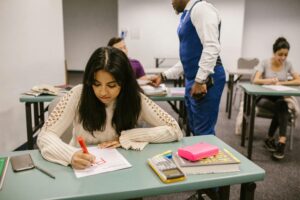 Female student writing on a paper at a desk in a classroom, with a teacher and other students in the background. | TCS