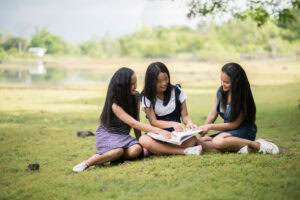 Three girls sitting on grass outside, smiling and reading a book together in a park-like setting. | TCS
